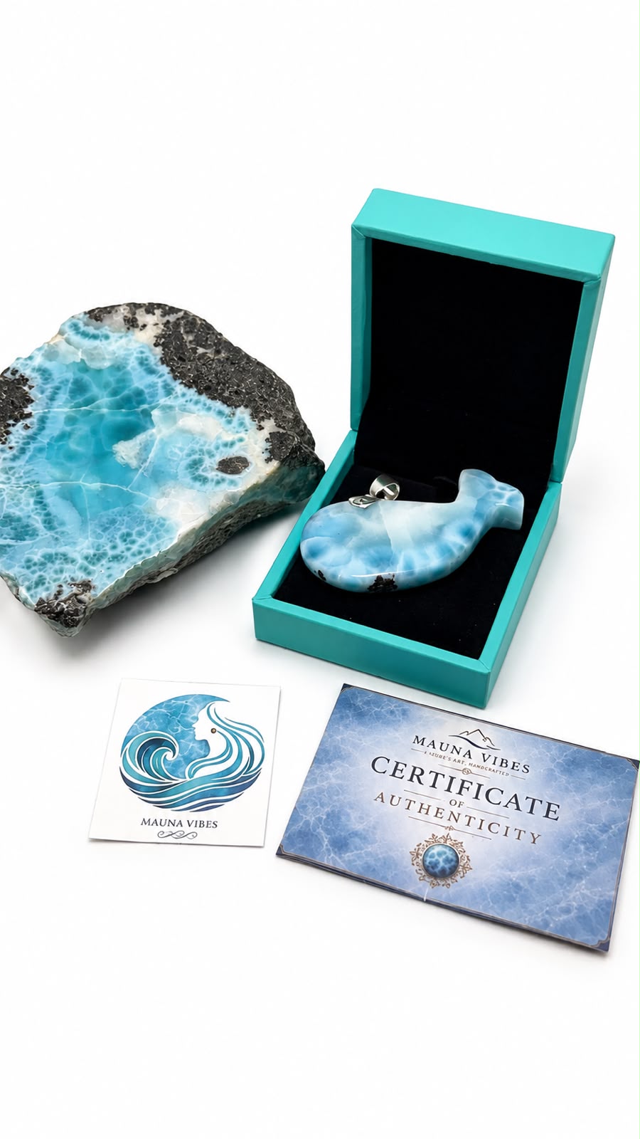 A larimar fish-shaped pendant displayed in an open turquoise box, alongside a large blue-and-black marbled stone, a small branded card, and a certificate card on a white background.