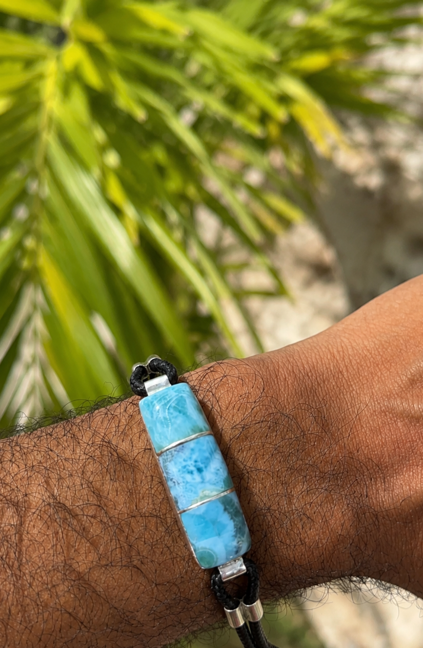 A close-up of a person's wrist wearing a lLarimar bracelet with a rectangular turquoise-blue stone centerpiece, black cord, and silver-toned metal accents, with blurred green foliage in the background.