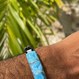 A close-up of a person's wrist wearing a lLarimar bracelet with a rectangular turquoise-blue stone centerpiece, black cord, and silver-toned metal accents, with blurred green foliage in the background.
