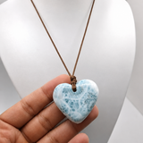 A heart-shaped Larimar made of light blue and white marbled stone, suspended from a brown cord necklace, held in a hand in front of a white mannequin bust.