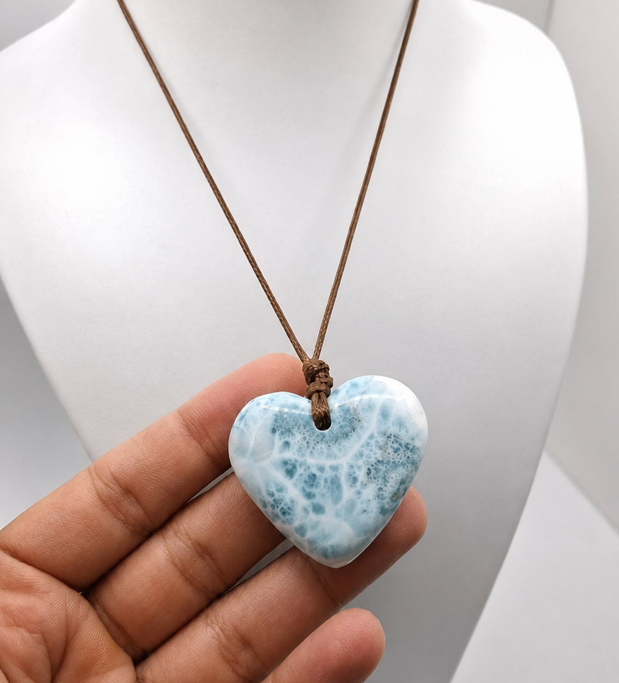A heart-shaped Larimar made of light blue and white marbled stone, suspended from a brown cord necklace, held in a hand in front of a white mannequin bust.