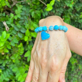 A turquoise blue beaded bracelet with a heart-shaped turquoise charm, shown worn on a person's wrist against a background of green foliage. A 'MAUNA UVI VIBES' logo is visible in the top-left corner.