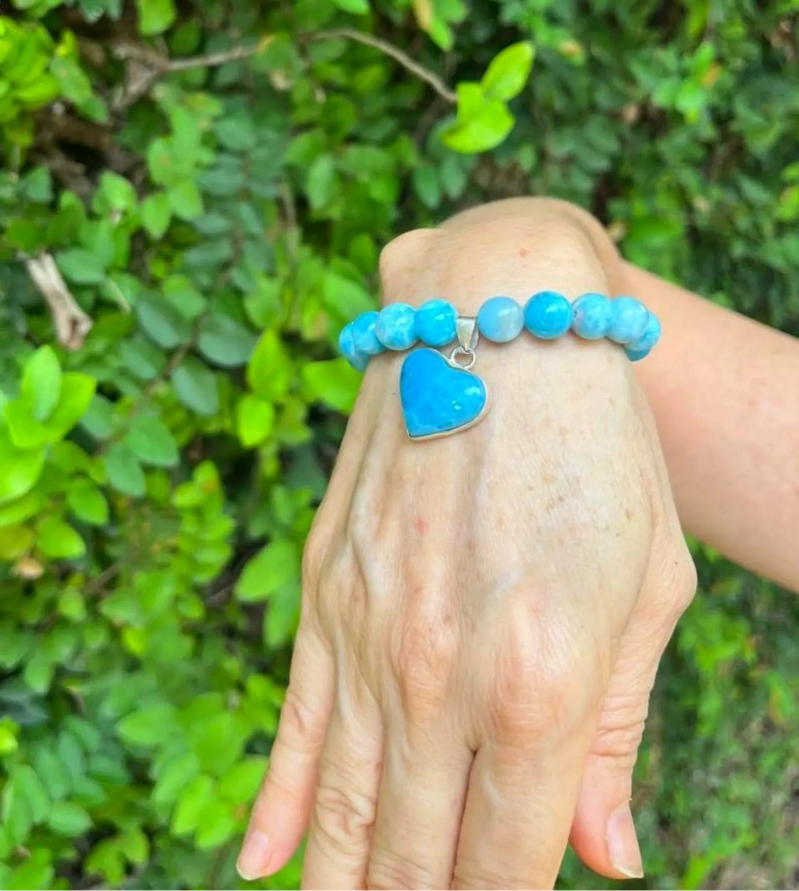 A turquoise blue beaded bracelet with a heart-shaped turquoise charm, shown worn on a person's wrist against a background of green foliage. A 'MAUNA UVI VIBES' logo is visible in the top-left corner.