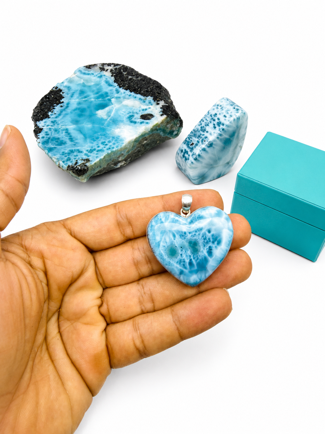 A heart-shaped Larimar stone pendant held in a person's hand, with two larger Larimar stone pieces and a turquoise box in the background on a white surface.