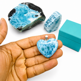 A heart-shaped Larimar stone pendant held in a person's hand, with two larger Larimar stone pieces and a turquoise box in the background on a white surface.