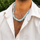 A man wearing a light blue Larimar  beaded necklace and a white open-collar shirt, photographed outdoors against a background of green trees.