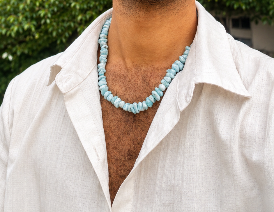 A man wearing a light blue Larimar  beaded necklace and a white open-collar shirt, photographed outdoors against a background of green trees.