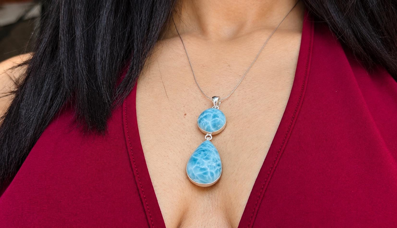 A close-up of a woman wearing a silver Larimar pendant necklace with two stacked Larimar stones, one oval and one teardrop, set against a deep red top.