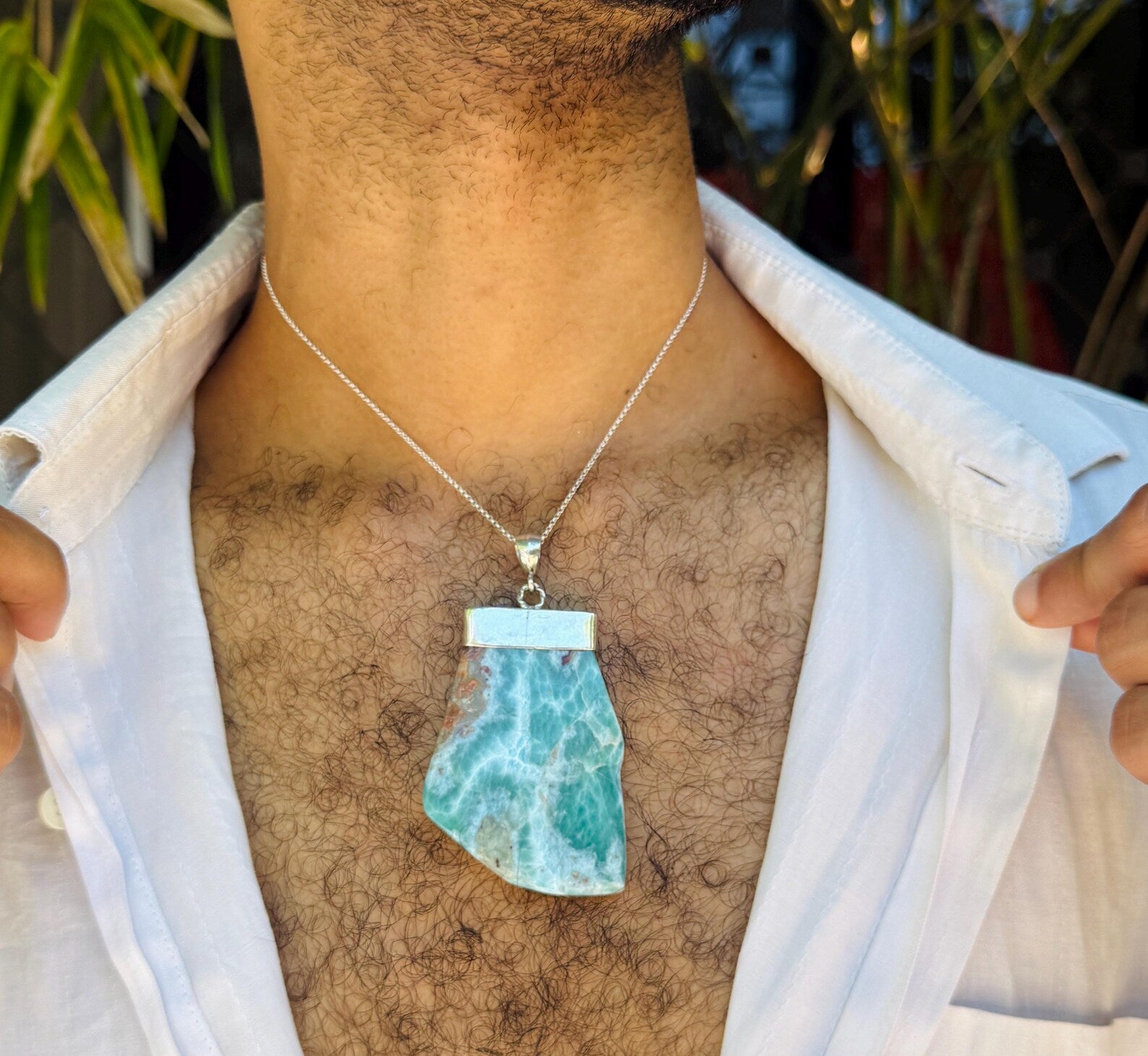 A close-up of a man wearing a silver chain necklace with a raw, irregular-shaped light blue and white marbled stone pendant, styled with an open white shirt.