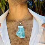 A close-up of a man wearing a silver chain necklace with a raw, irregular-shaped light blue and white marbled stone pendant, styled with an open white shirt.