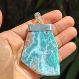 A hand holding a raw, polished Larimar pendant with a silver-toned metal cap and bail, set against a blurred background of green foliage.