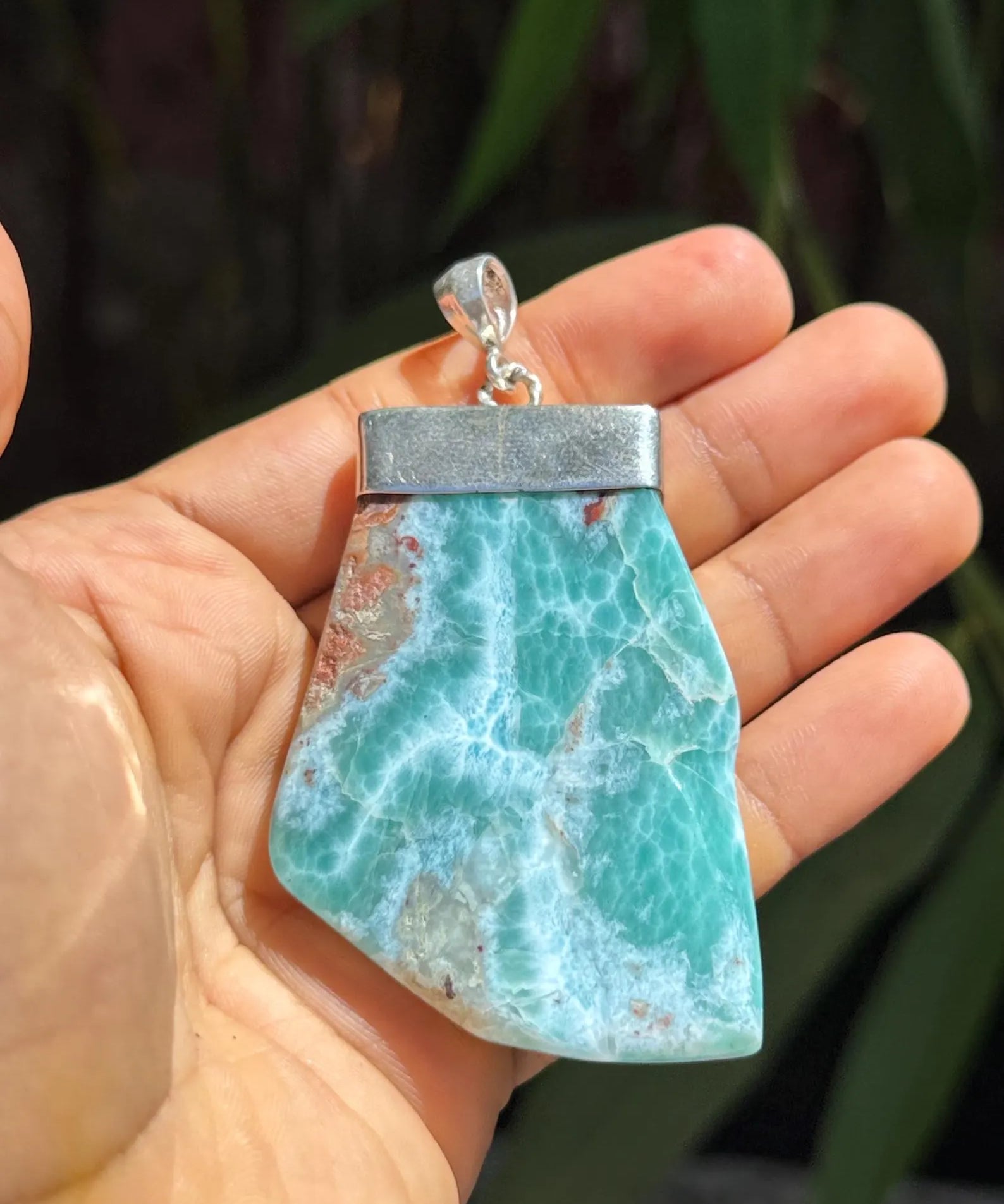 A hand holding a raw, polished Larimar pendant with a silver-toned metal cap and bail, set against a blurred background of green foliage.
