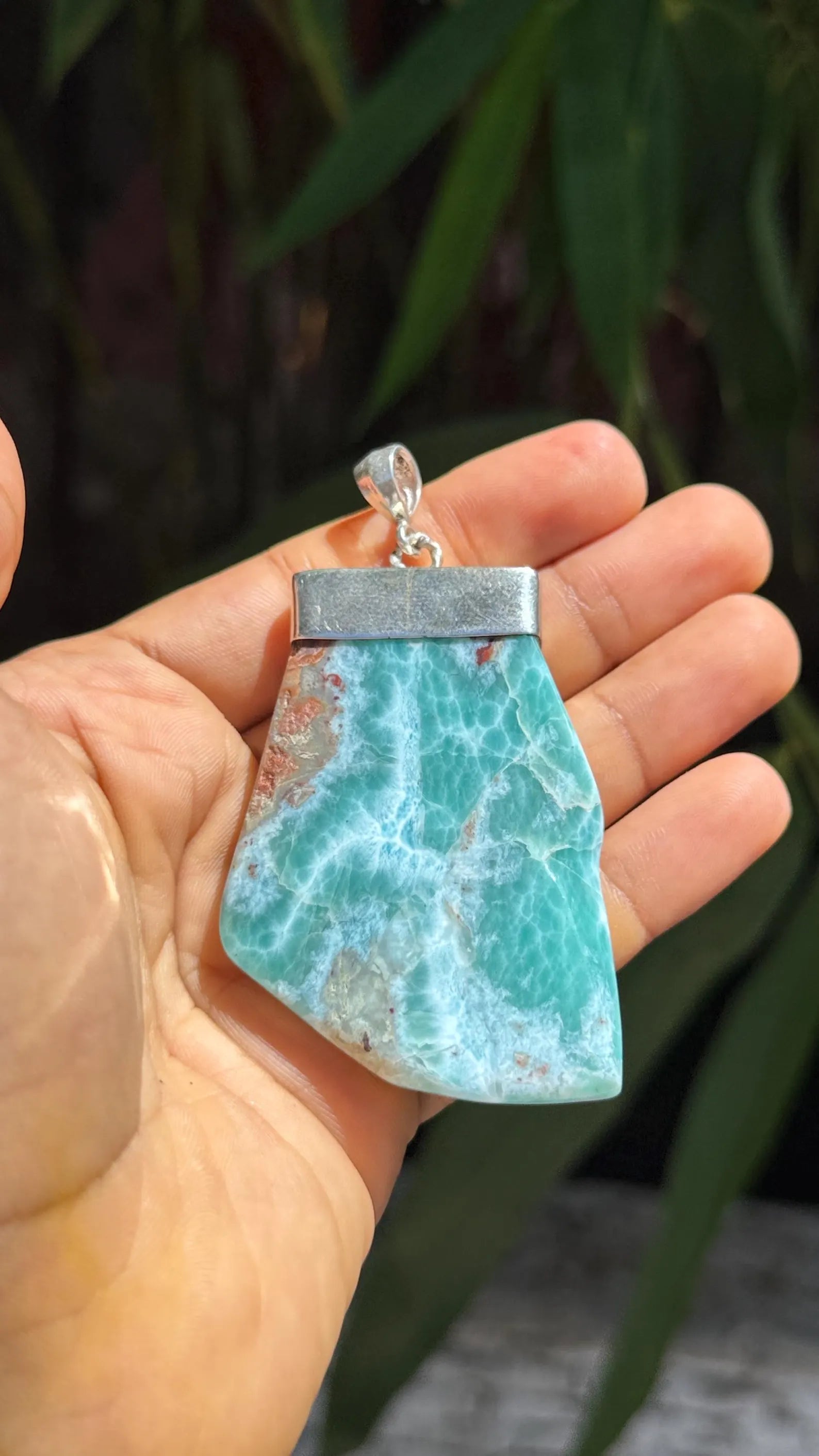 A hand holding a raw, polished Larimar pendant with a silver-toned metal cap and bail, set against a blurred background of green foliage.