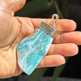 A hand holding a raw larimar stone pendant with a silver metal cap and loop, set against a blurred background of green leaves.