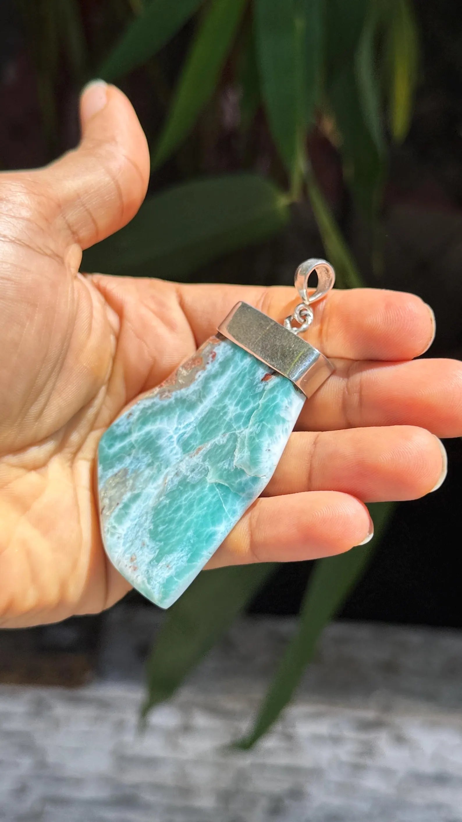 A hand holding a raw larimar stone pendant with a silver metal cap and loop, set against a blurred background of green leaves.