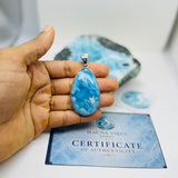 A hand holds a teardrop-shaped blue stone pendant with a silver bail, positioned above a Certificate of Authenticity card and a larger blue stone specimen. The pendant features marbled blue and white patterns.