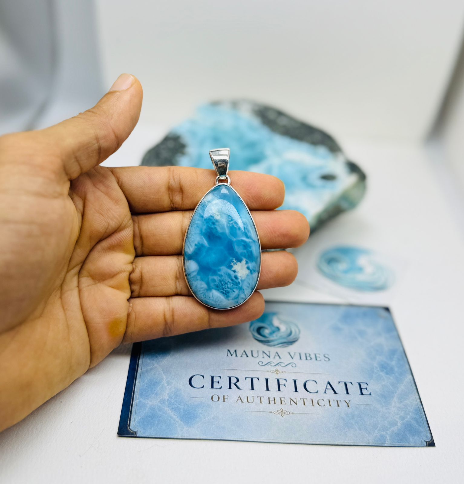 A hand holds a teardrop-shaped blue stone pendant with a silver bail, positioned above a Certificate of Authenticity card and a larger blue stone specimen. The pendant features marbled blue and white patterns.