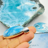 A hand holds a silver ring with a polished cabochon gemstone featuring a swirling light blue and white marbled pattern. In the background, a larger matching stone slab and a certificate card are visible on a white surface.