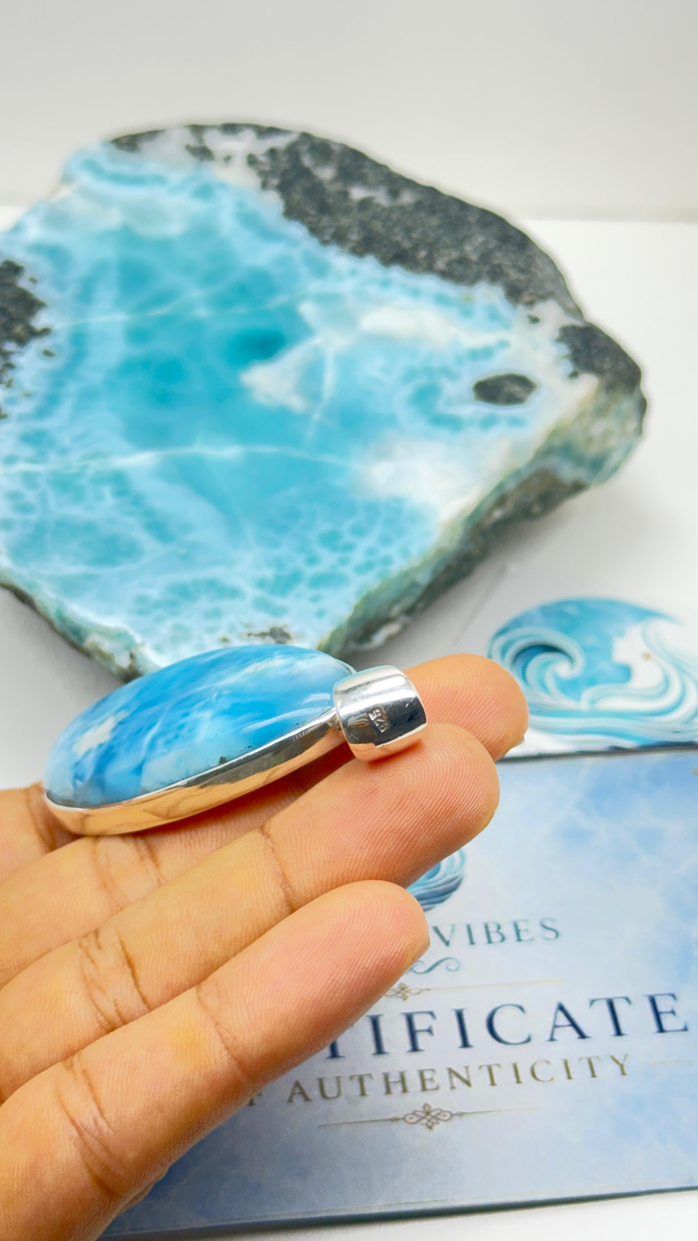 A hand holds a silver ring with a polished cabochon gemstone featuring a swirling light blue and white marbled pattern. In the background, a larger matching stone slab and a certificate card are visible on a white surface.