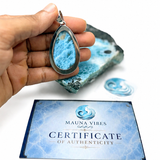 A hand holds a teardrop-shaped pendant made from Larimar stone in a silver bezel, with a matching blue stone slab and a blue 'Certificate of Authenticity' card from Mauna Vibes in the background on a white surface.