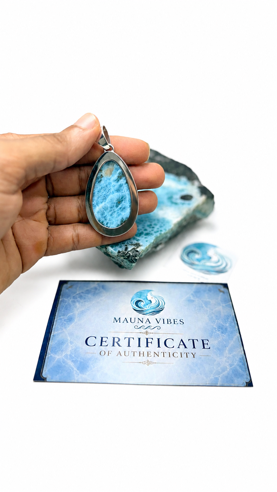 A hand holds a teardrop-shaped pendant made from Larimar stone in a silver bezel, with a matching blue stone slab and a blue 'Certificate of Authenticity' card from Mauna Vibes in the background on a white surface.