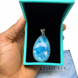 A hand holds an open turquoise jewelry box containing a teardrop-shaped blue marbled pendant with a silver bail. A certificate card is visible underneath on a white background.