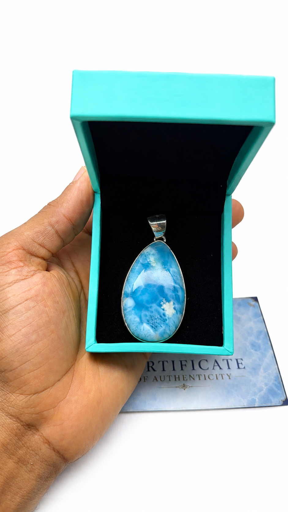A hand holds an open turquoise jewelry box containing a teardrop-shaped blue marbled pendant with a silver bail. A certificate card is visible underneath on a white background.