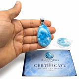A hand holds a teardrop-shaped blue and white marbled pendant with a silver bail, positioned above a blue 'Certificate of Authenticity' card from Mauna Vibes. A small matching blue stone is visible in its packaging in the background, all on a clean white studio background.