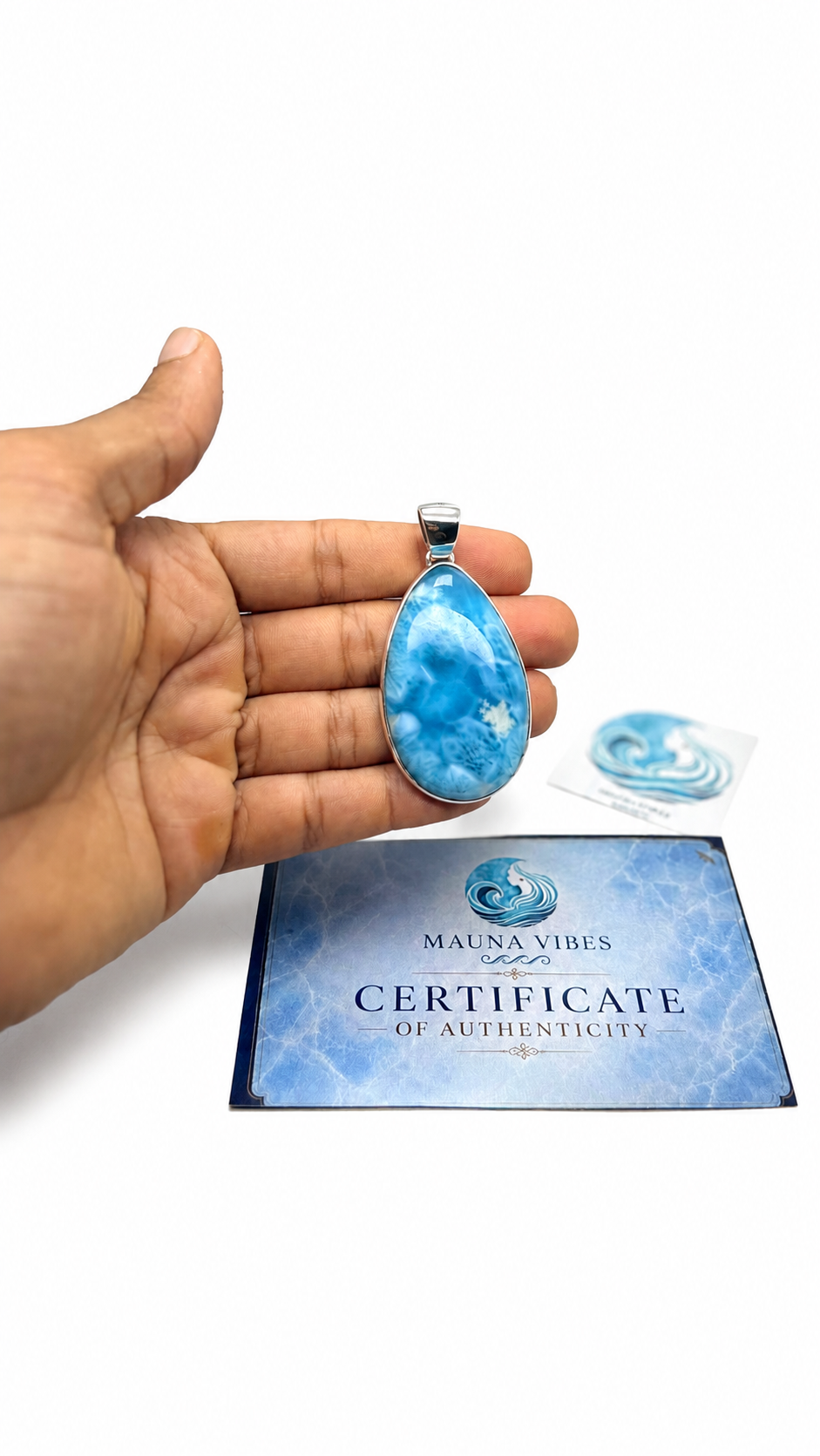 A hand holds a teardrop-shaped blue and white marbled pendant with a silver bail, positioned above a blue 'Certificate of Authenticity' card from Mauna Vibes. A small matching blue stone is visible in its packaging in the background, all on a clean white studio background.
