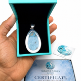 A hand holds an open turquoise jewelry box displaying a teardrop-shaped blue stone pendant with a silver bezel. A small branded card and a Certificate of Authenticity from Mauna Vibes are placed below on a white background.