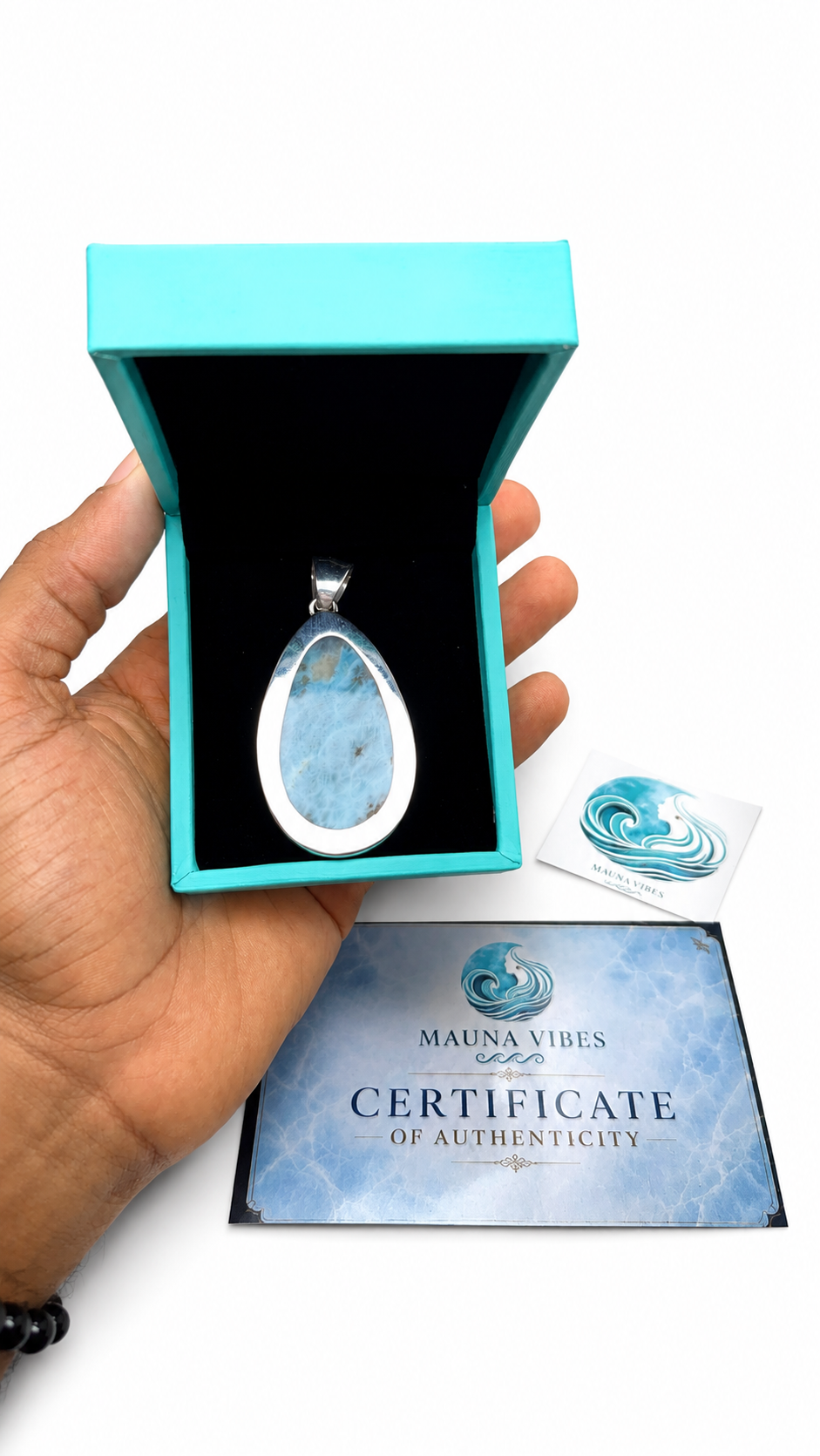 A hand holds an open turquoise jewelry box displaying a teardrop-shaped blue stone pendant with a silver bezel. A small branded card and a Certificate of Authenticity from Mauna Vibes are placed below on a white background.