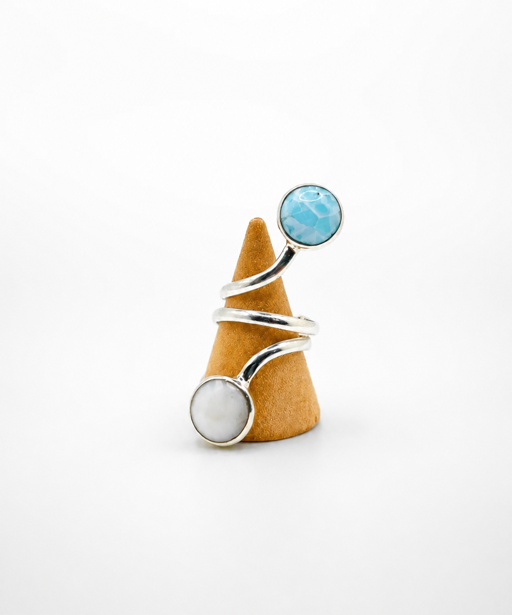 A silver wrap ring with a round light blue stone on one end and a round white stone on the other, displayed on a small cone-shaped stand against a white background.