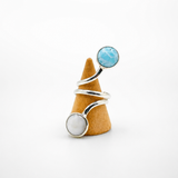 A silver wrap ring with a round light blue stone on one end and a round white stone on the other, displayed on a small cone-shaped stand against a white background.