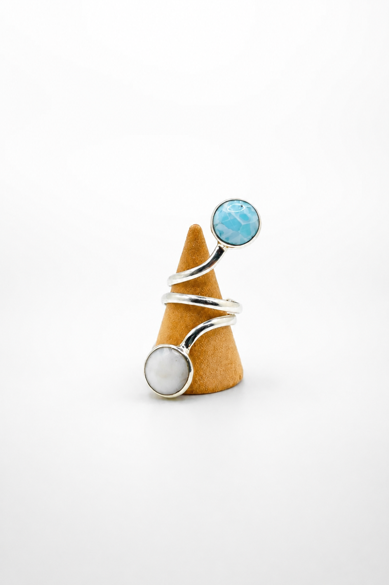 A silver wrap ring with a round light blue stone on one end and a round white stone on the other, displayed on a small cone-shaped stand against a white background.
