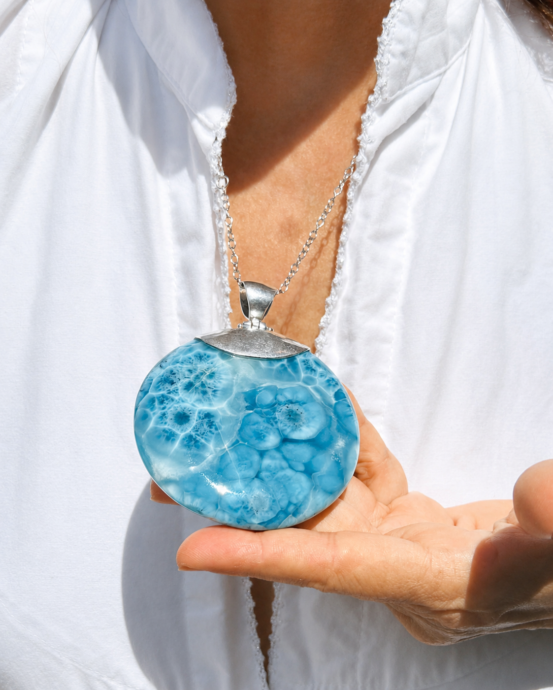 A woman wearing a round Larimar Pendant in a white blouse holds a round pendant necklace with a silver-toned cap and chain. The pendant is light blue with darker blue marbled or bubble-like patterning.