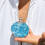 A woman wearing a round Larimar Pendant in a white blouse holds a round pendant necklace with a silver-toned cap and chain. The pendant is light blue with darker blue marbled or bubble-like patterning.
