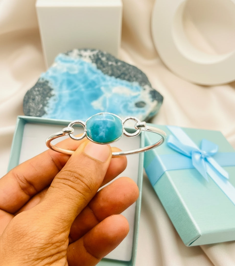 Hand holding a silver bracelet with a turquoise stone in front of a gift box and heart-shaped object.