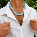 A man wearing a larimar  beaded necklace and a white button-down shirt, photographed outdoors with green foliage in the background.