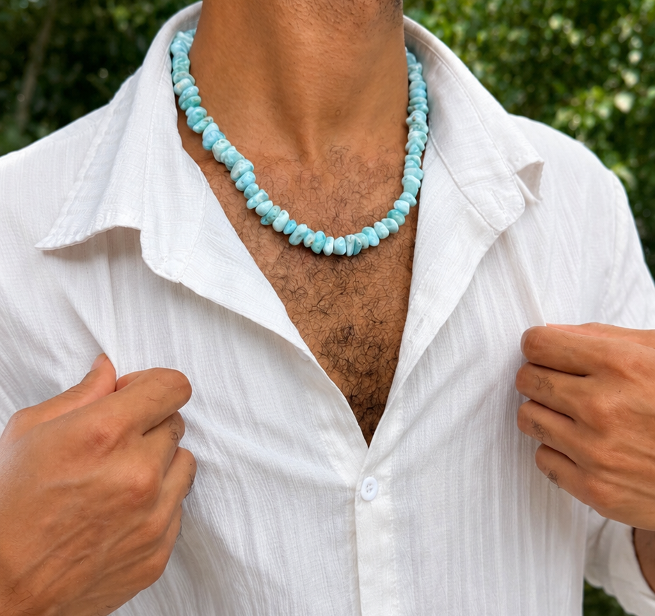 A man wearing a larimar  beaded necklace and a white button-down shirt, photographed outdoors with green foliage in the background.