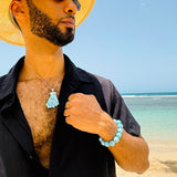 A man on a beach wearing a straw hat, black shirt, and sunglasses, showcasing a light blue tassel pendant necklace and a matching light blue beaded bracelet.