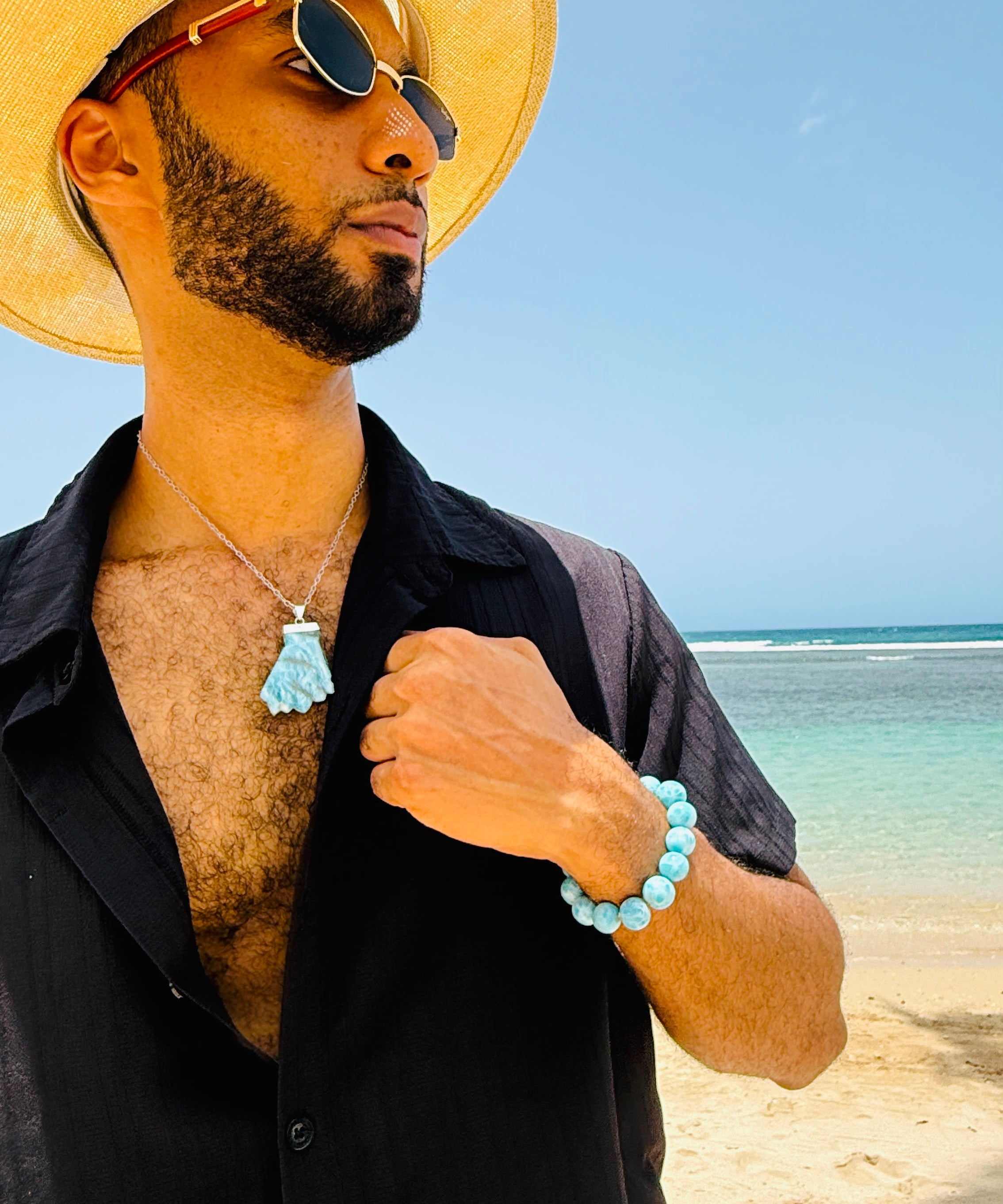 A man on a beach wearing a straw hat, black shirt, and sunglasses, showcasing a light blue tassel pendant necklace and a matching light blue beaded bracelet.
