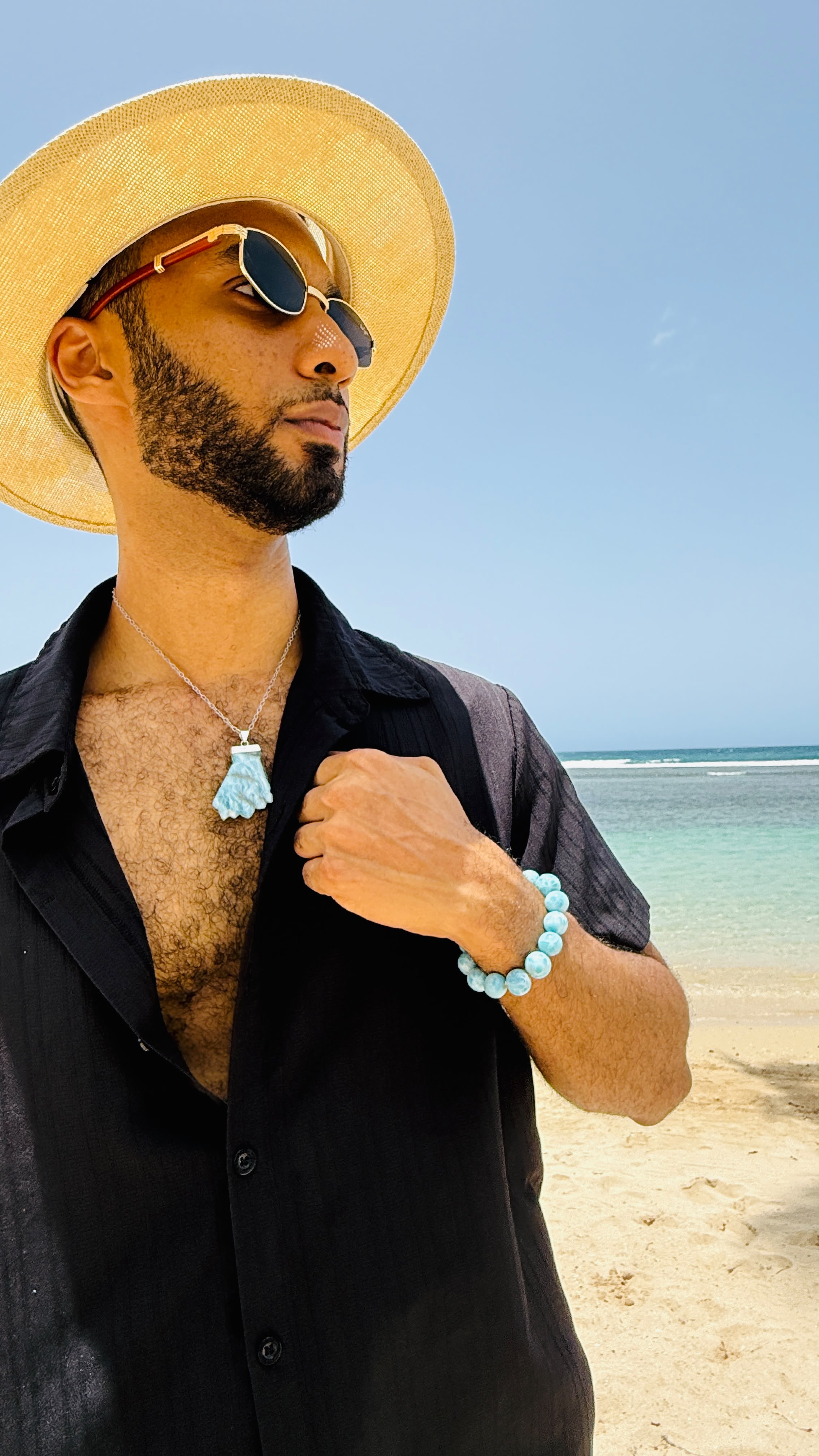 A man on a beach wearing a straw hat, black shirt, and sunglasses, showcasing a light blue tassel pendant necklace and a matching light blue beaded bracelet.