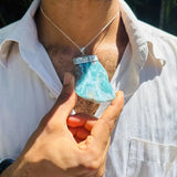 A person wearing a silver pendant necklace with a larimar stone pendant, holding the pendant closer to the camera with one hand.