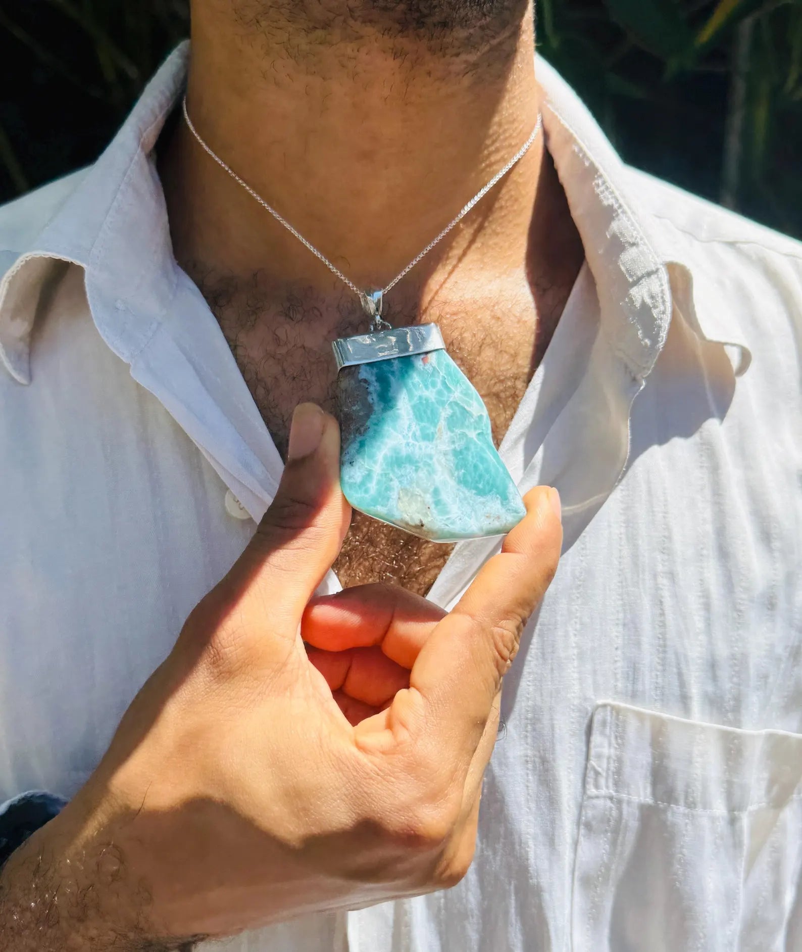 A person wearing a silver pendant necklace with a larimar stone pendant, holding the pendant closer to the camera with one hand.