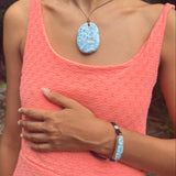 A woman wearing a coral pink textured dress and a light pink headwrap, showcasing a Larimar stone pendant necklace and a matching light blue beaded bracelet.