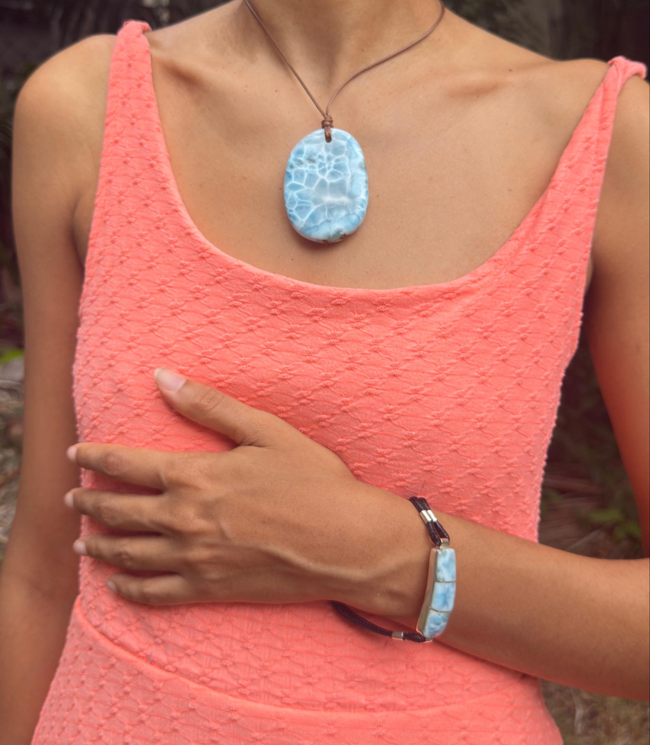 A woman wearing a coral pink textured dress and a light pink headwrap, showcasing a Larimar stone pendant necklace and a matching light blue beaded bracelet.