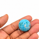 A round turquoise-blue stone bead with a small silver jump ring, held in the palm of a hand against a plain white background.