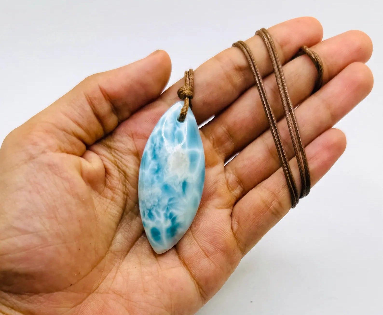 A hand holding a teardrop-shaped Larimar pendant necklace with a brown cord, set against a plain white background.