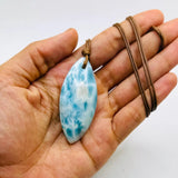 A hand holding a teardrop-shaped Larimar pendant necklace with a brown cord, set against a plain white background.
