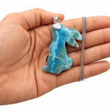 A hand holding a carved blue  Larimar and white marbled pendant on a silver chain, shown against a plain white background.